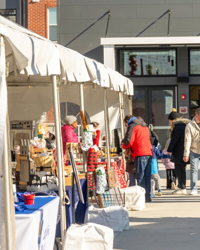 Shoppers browsing holiday vendor booths under white tents on an outdoor plaza.