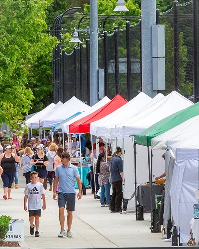 Shoppers walking past white and colored market tents on a tree-lined street in Woodinville