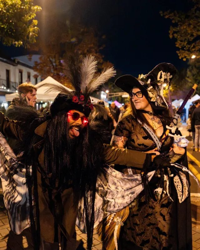Two costumed revelers in feathered hats and red sunglasses on an evening street at the Halloween Night Market in Haddonfield.