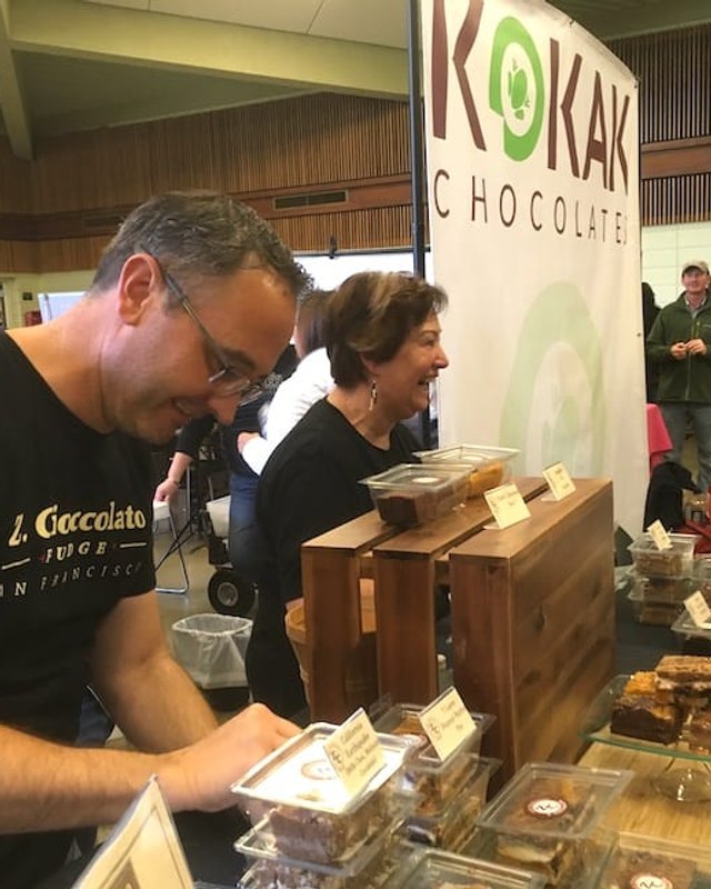 A man in a black Cioccolato t-shirt and a smiling woman at the KOKAK Chocolate booth, with people in the hall behind them.