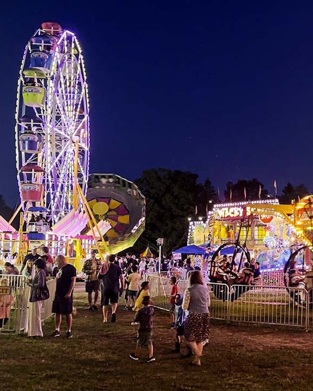 Night crowd at a lit-up fair with a Ferris wheel and illuminated ride booths.