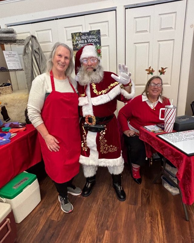 Santa and two women, one in a red apron, at a holiday bazaar table inside a church hall, with red table linens and a card display.