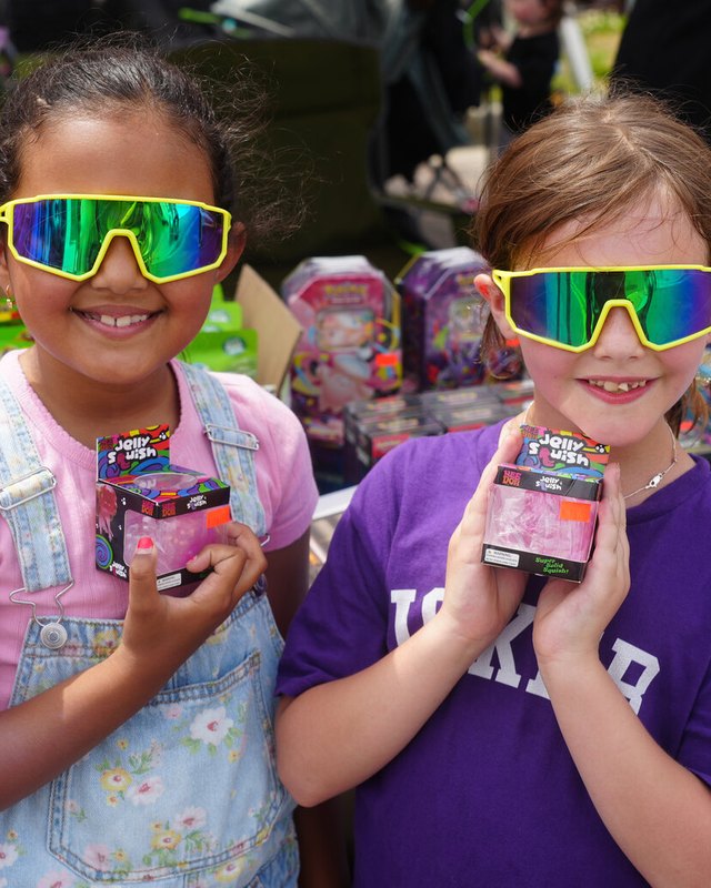 Two girls in rainbow sunglasses holding prizes in front of a festival game booth stocked with toys.