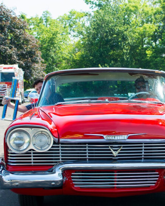Red classic car with a chrome grille on display at an outdoor show on a sunny day, with trees and people in the background.