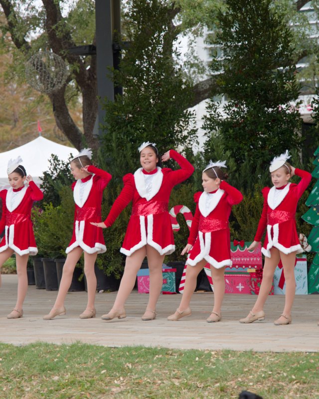Dancers in red and white holiday outfits performing on an outdoor stage at Levy Park, with white tents and evergreen trees behind them.