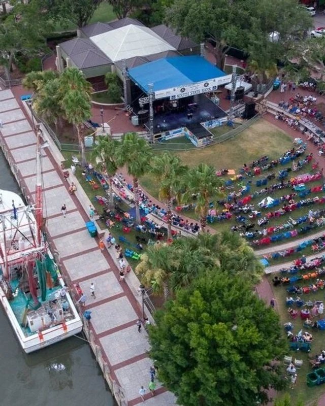 Aerial view of a waterfront festival crowd gathered around a stage with tents and a docked boat.