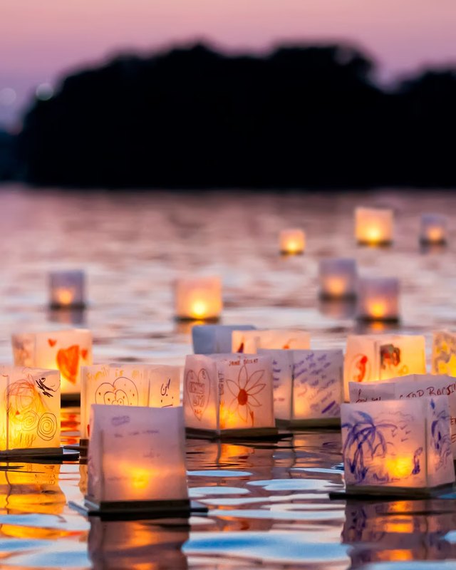 Floating paper lanterns with drawings and candles glowing on calm water at sunset.