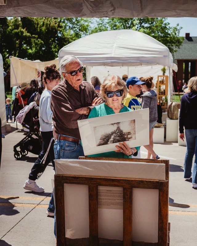 Two shoppers look at a framed artwork at an outdoor booth at the Marion Arts Festival, with white tents and crowds around them.