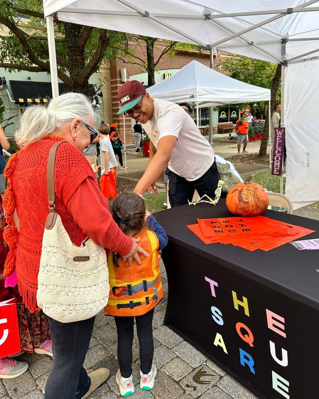 A grandmother and a young child in an orange vest at a The Square booth with a pumpkin and orange sheets on a black table outdoors.