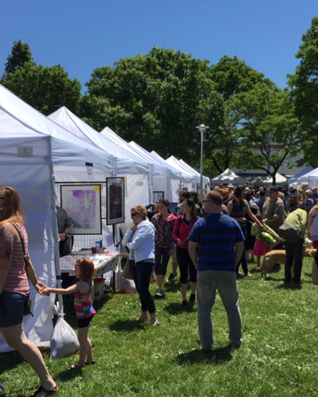 Crowd of shoppers walking between white booth tents at an outdoor craft fair on a sunny day.