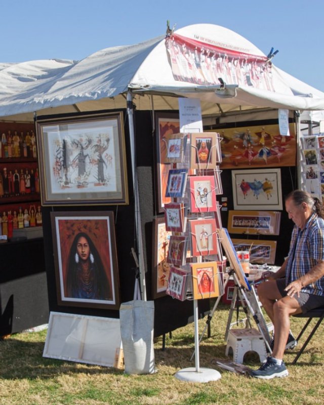 Art vendor booth with framed paintings and prints under a white tent at an outdoor festival, with a man seated nearby.