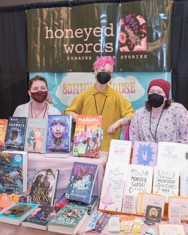 Vendor booth with stacked books and zines behind a Honeyed Words banner, with three people staffing the table.