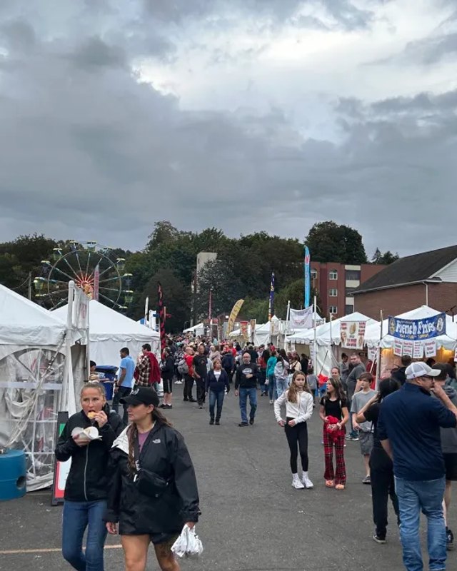 Crowd of visitors walking past white vendor tents at an outdoor autumn festival on a cloudy day.