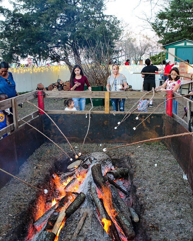 People roast marshmallows on sticks over a log fire pit at Christmas in the Park in Mesquite, with holiday lights behind a fence.