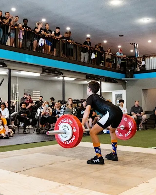 Weightlifter lifting a barbell in front of a crowd watching from a gym balcony and floor seating.