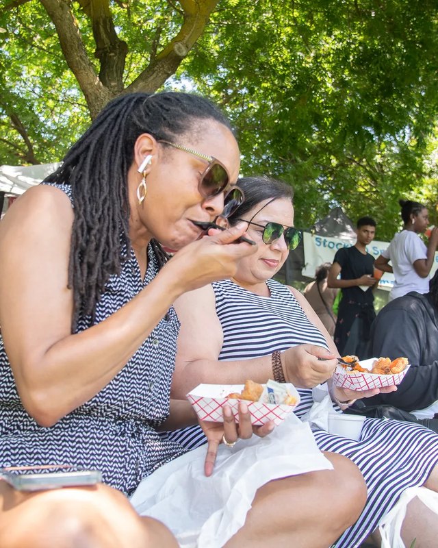 Two women in striped dresses eating fried food from red-and-white paper trays at an outdoor Juneteenth celebration, with trees and a crowd behind them.