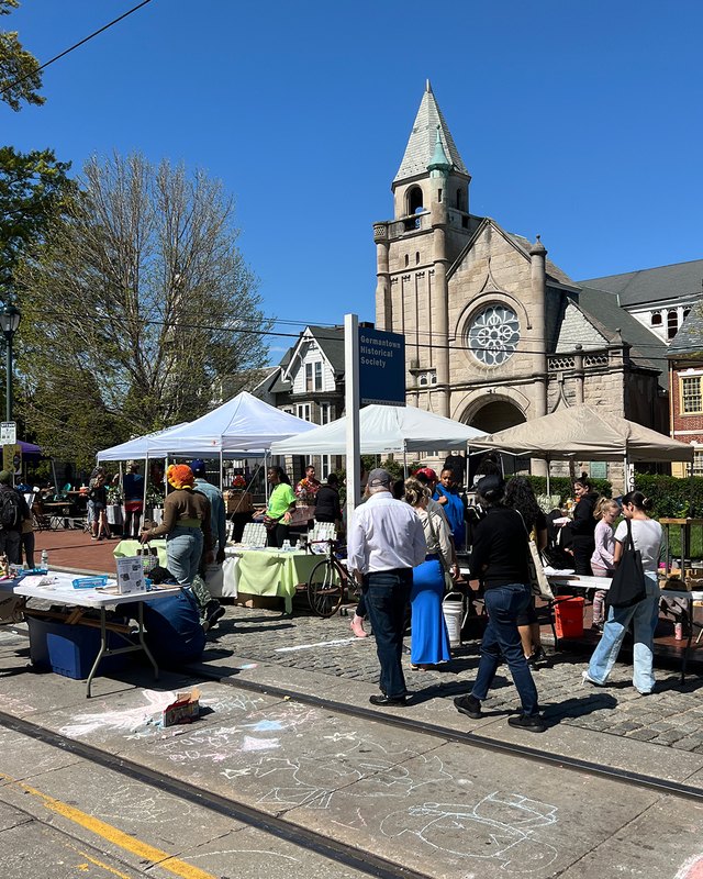 Market stalls with tents and shoppers on the square in front of a stone church on a sunny day.