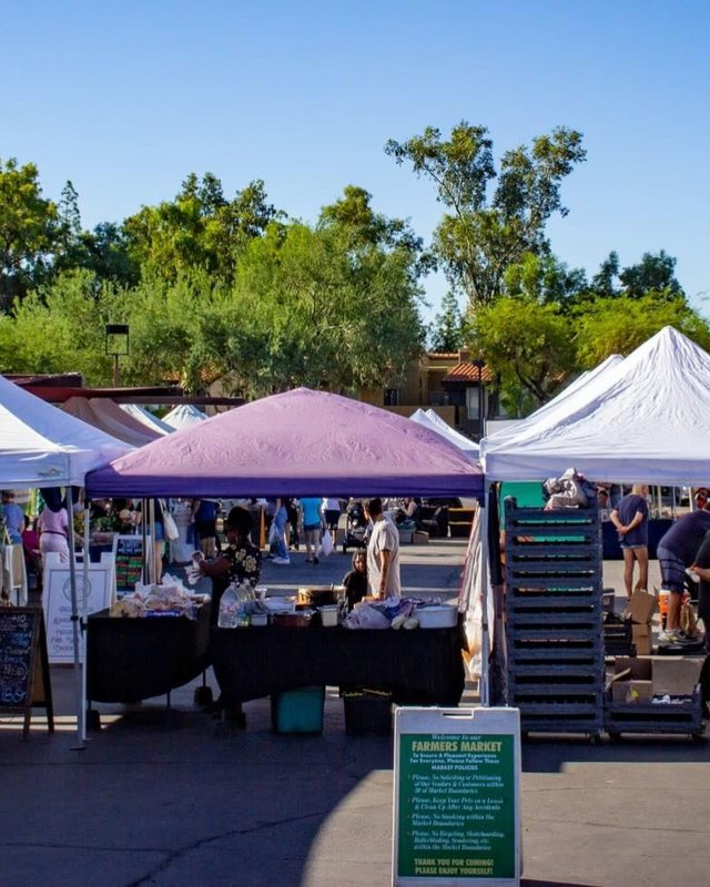 Shoppers browsing white-tent market booths at the Ahwatukee Farmers Market under a clear blue sky.