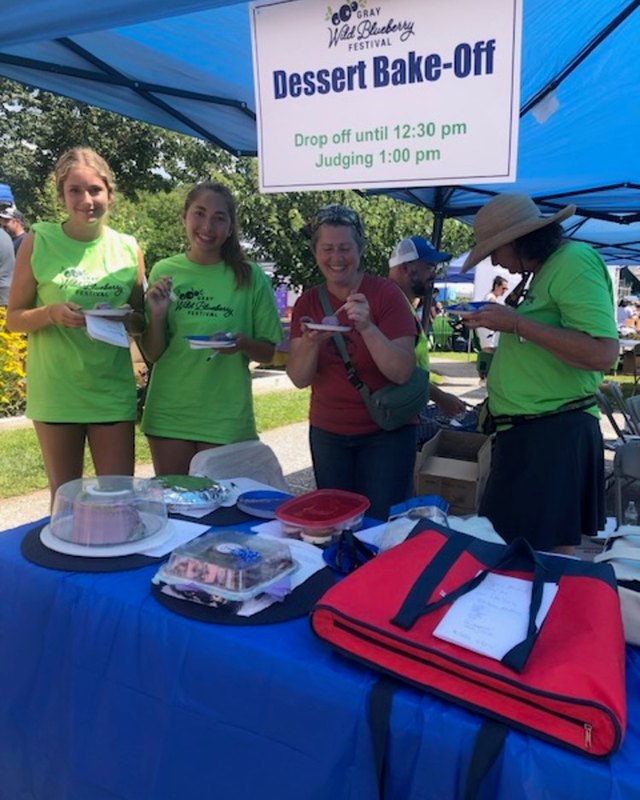 Volunteers in green shirts at a Dessert Bake-Off table under a blue tent at the Gray Wild Blueberry Festival.