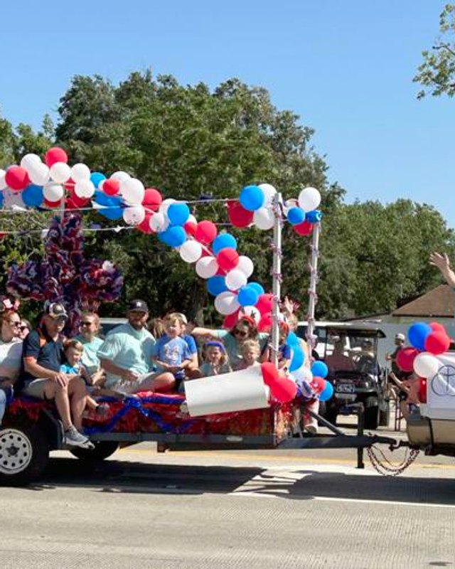 Parade float decorated with red, white and blue balloons, carrying riders on a flatbed trailer; a truck behind bears a Columbia Methodist Church banner.