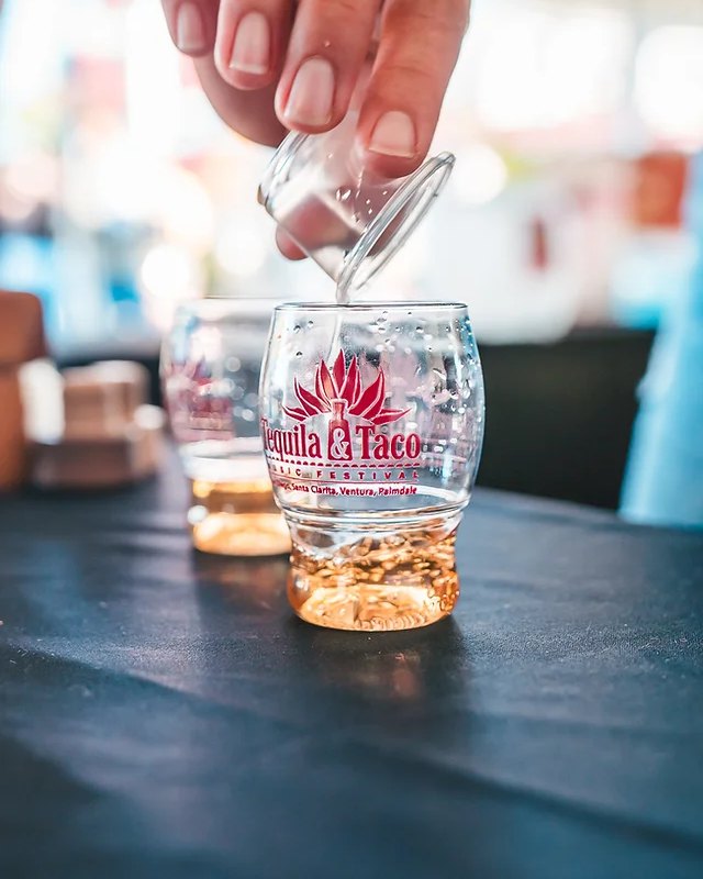 Shot glasses with the Tequila & Taco logo being poured on a dark bar table at the festival.