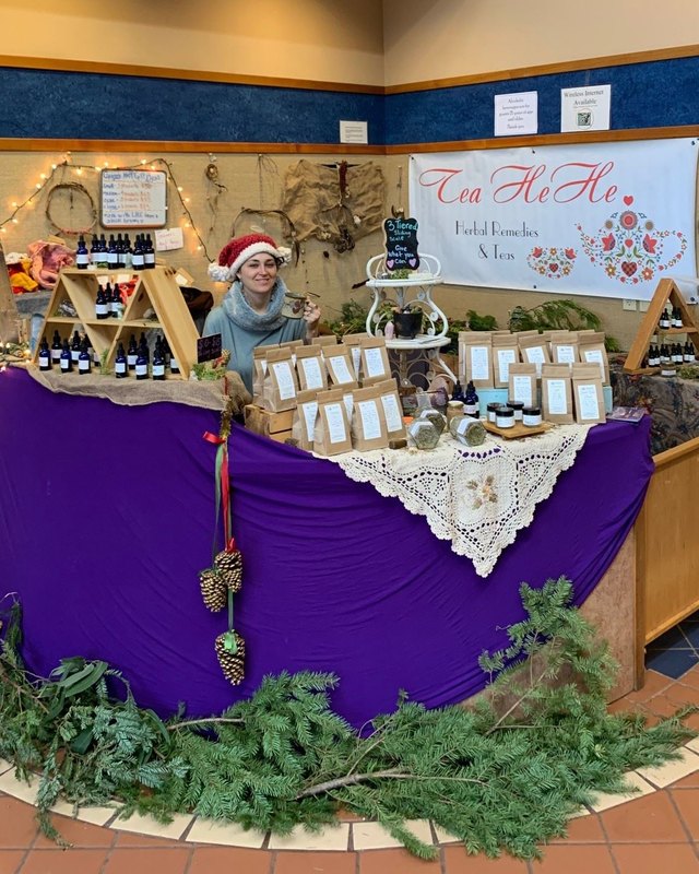 A holiday craft booth with jars and goods on a purple tablecloth, with a vendor in a Santa hat behind it