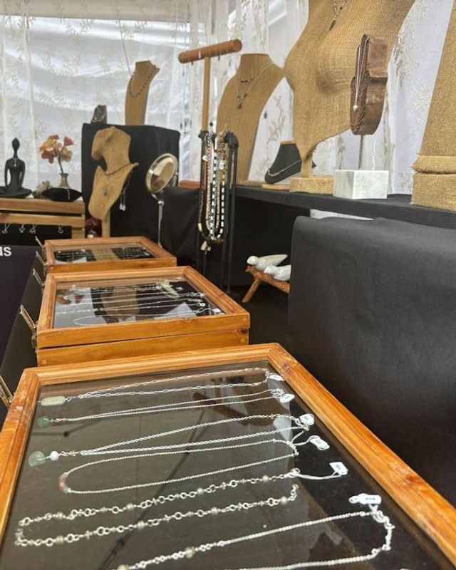 Jewelry necklaces laid out in wooden-framed glass cases on black tables at an arts fair booth, with mannequin busts and linen displays behind.
