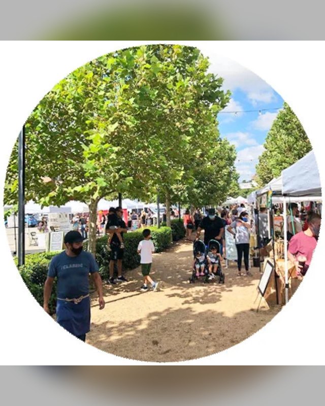 Shoppers and vendor tents along a tree-lined path at an outdoor market.