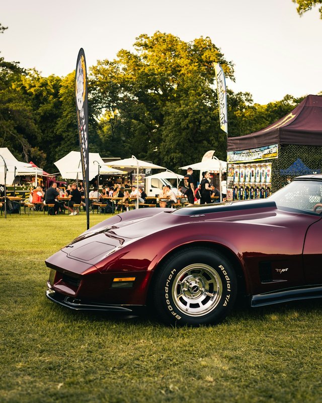 Classic burgundy Corvette parked on grass beside tents and shoppers at an outdoor car show.