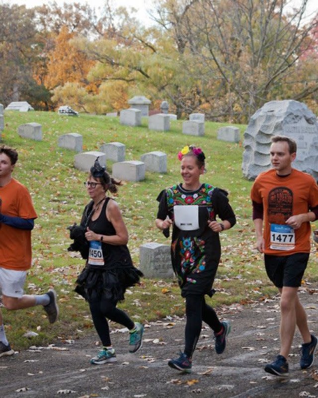 Runners in orange shirts and costumes jogging past old headstones in an autumn cemetery.