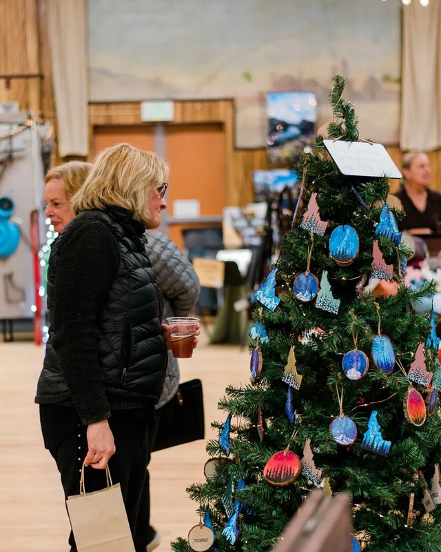 Shopper holding a drink beside a decorated tree with ornaments, with craft vendor booths behind her at the holiday market.