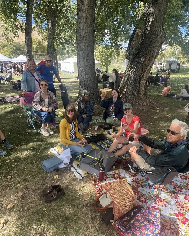 Groups of people relaxing on blankets and lawn chairs under trees beside white vendor tents at an outdoor event.