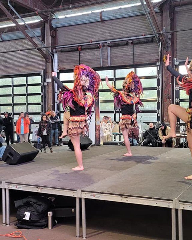 Dancers in colorful costumes performing on a stage in front of a crowd at an indoor pier venue.