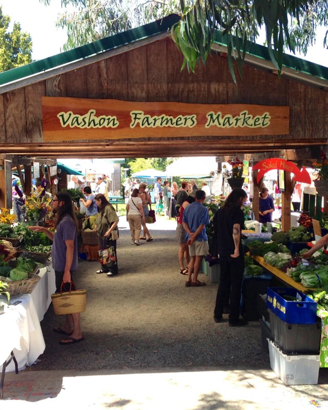 Shoppers and vendor booths under the Vashon Farmers Market sign at the village green market.