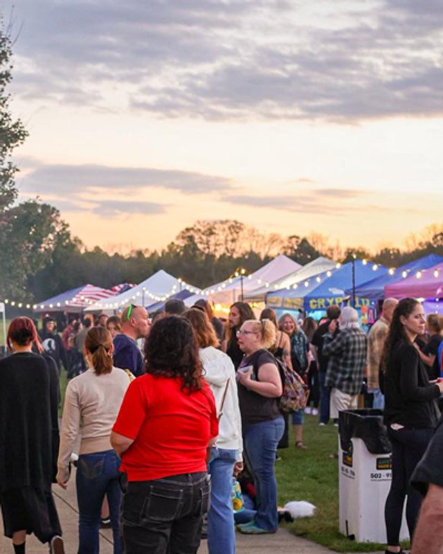 Shoppers and visitors walking past white vendor tents at an evening outdoor market