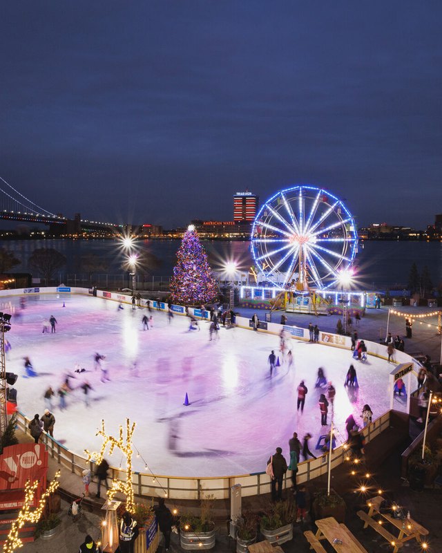 Skaters on the lit RiverRink ice rink with a Ferris wheel and bridge lights at dusk