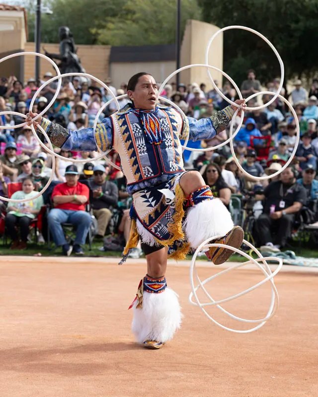 Hoop dancer in regalia performing with white hoops before a large outdoor crowd.
