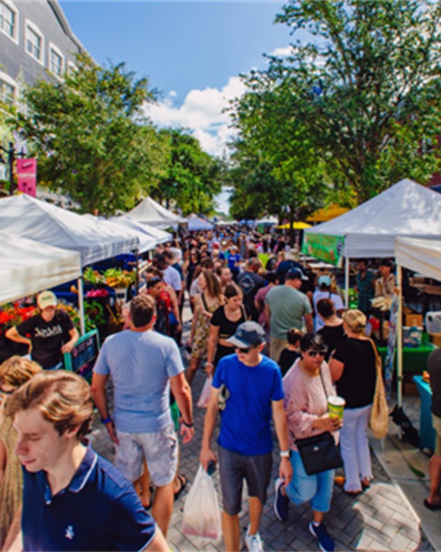 Shoppers walking between white-tent vendor booths at an outdoor market on a sunny day.