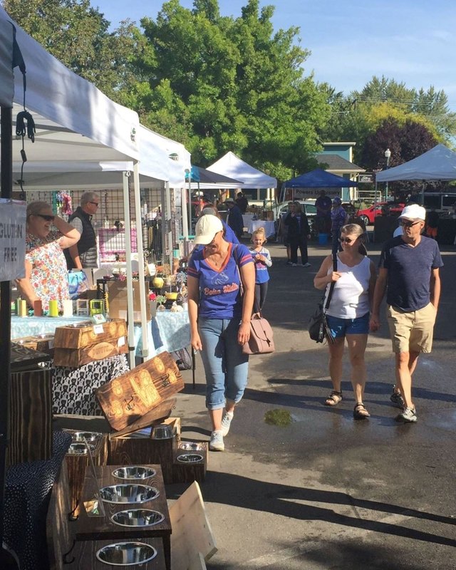 Shoppers walk past white-tent market booths with wooden displays and goods on tables at an outdoor market.