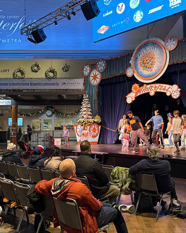 Winterfest stage with performers and a seated audience under holiday decorations at Seattle Center