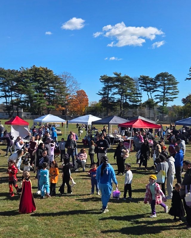 Crowd of costumed visitors on a grassy field among white and red vendor tents under a blue sky at an outdoor fall festival