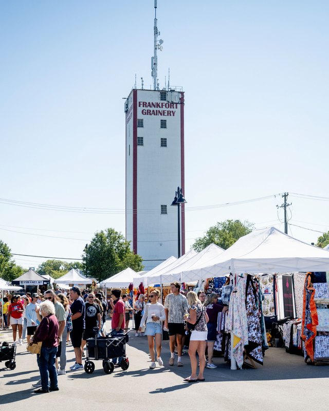 Shoppers walking past white vendor tents and clothing racks beneath the Frankfort Grainery tower at the fall festival.