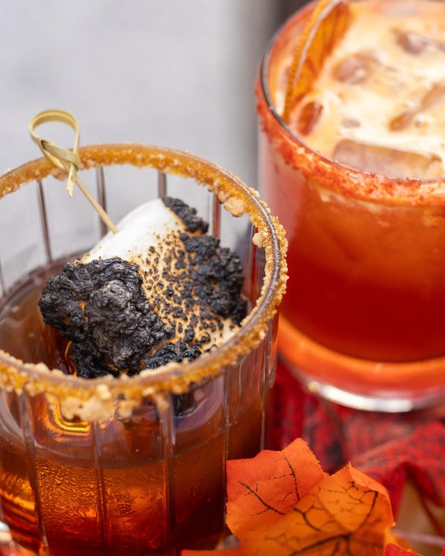 Two Halloween-themed cocktails with a torched smoked marshmallow garnish and orange autumn leaves on a red plaid cloth.