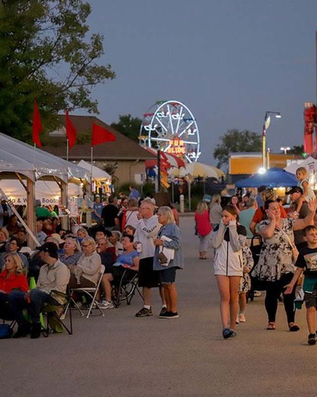 Evening crowd walking past white vendor tents and booths at the Manchester Homecoming midway.