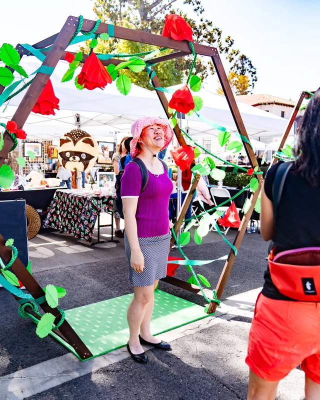 A woman in a pink hat stands beside a decorated wooden frame with fabric leaves and flowers, with market tents and vendor booths behind her.