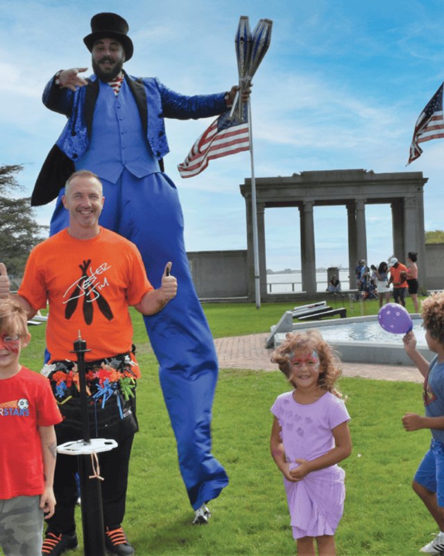 Stilt walker and children posing on a lawn at a park event with tents and flags nearby.