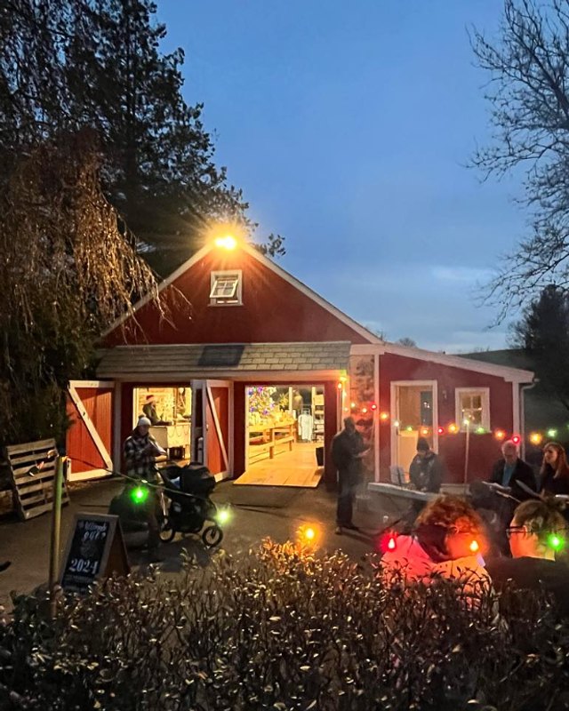 Evening view of the lit red barn and farm buildings at Spring Creek Farm with string lights and visitors outside.