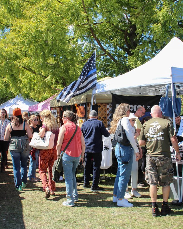 Shoppers browsing tents and booths at an outdoor fair under shady trees.