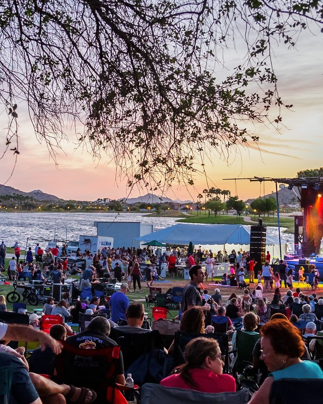 Crowd gathered on lawn chairs facing the lit stage at the Goodyear lakeside amphitheater at sunset.