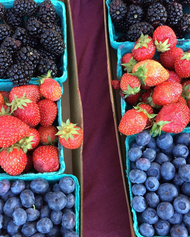 Cartons of fresh strawberries, blackberries and blueberries set out on a purple cloth at a market.